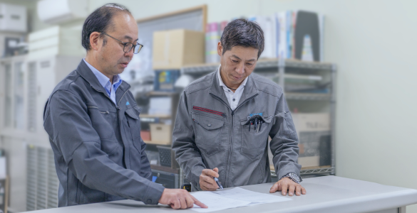 屋内の作業場にて、ヘルメットを着用したベテラン技術者が資材を前にして真剣な表情で確認・作業を行っている様子