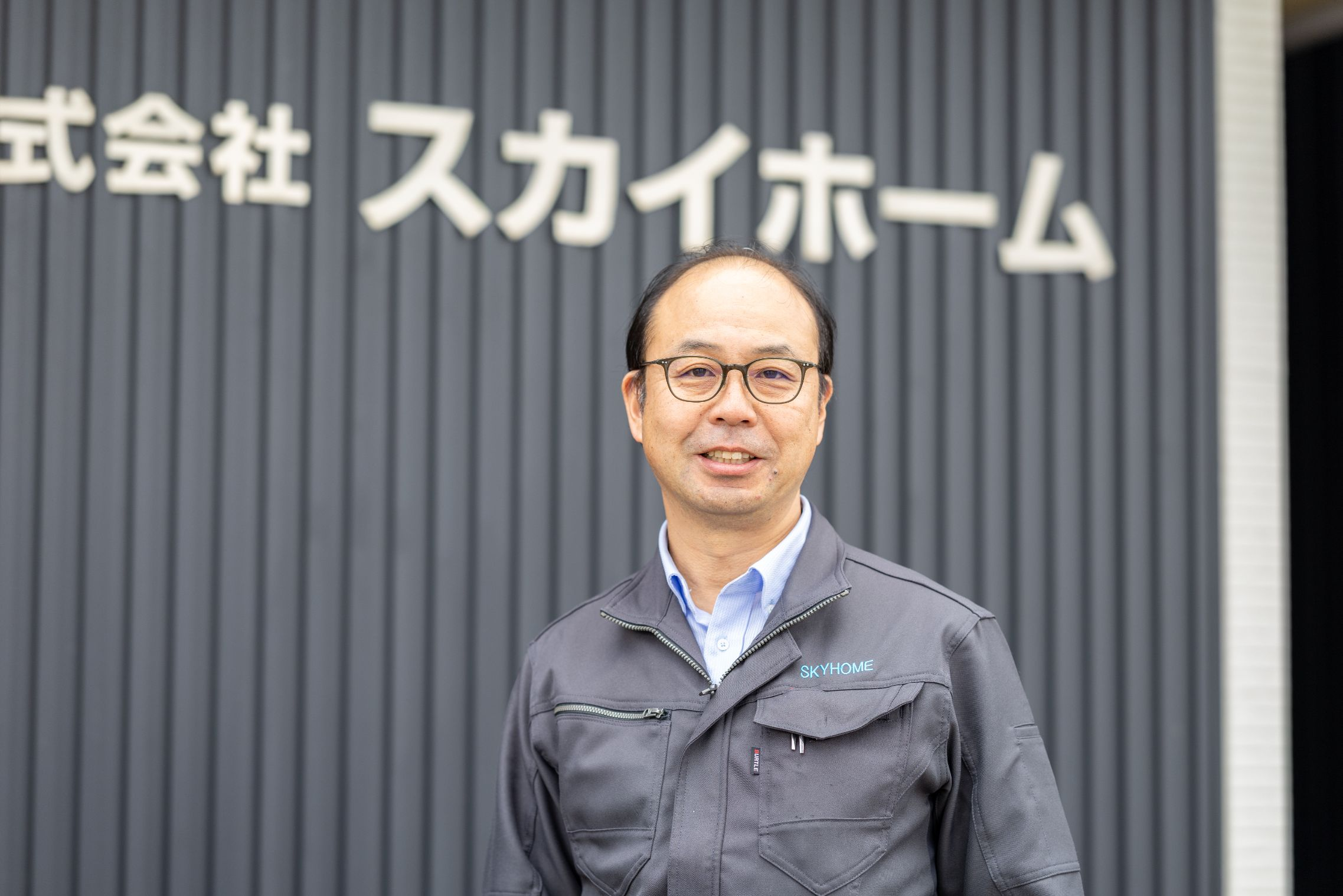 「株式会社スカイホーム」の看板を背景に、作業着姿で穏やかに微笑む代表取締役社長・河村 康平のポートレート