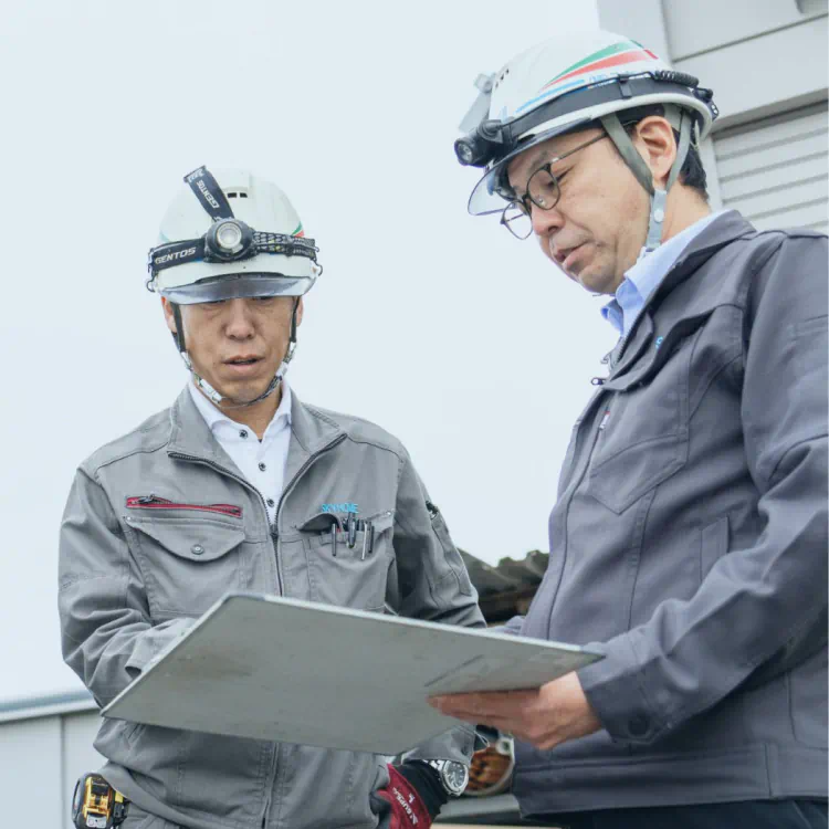 未経験からの育成：屋外で図面を広げ、社長が後輩に優しくアドバイスをしている様子