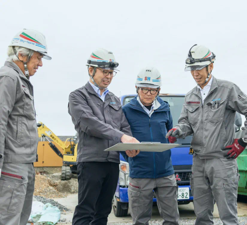 屋外の現場で、ヘルメットを着用した4人の社員たちがタブレットを囲んで施工管理の打ち合わせをする様子